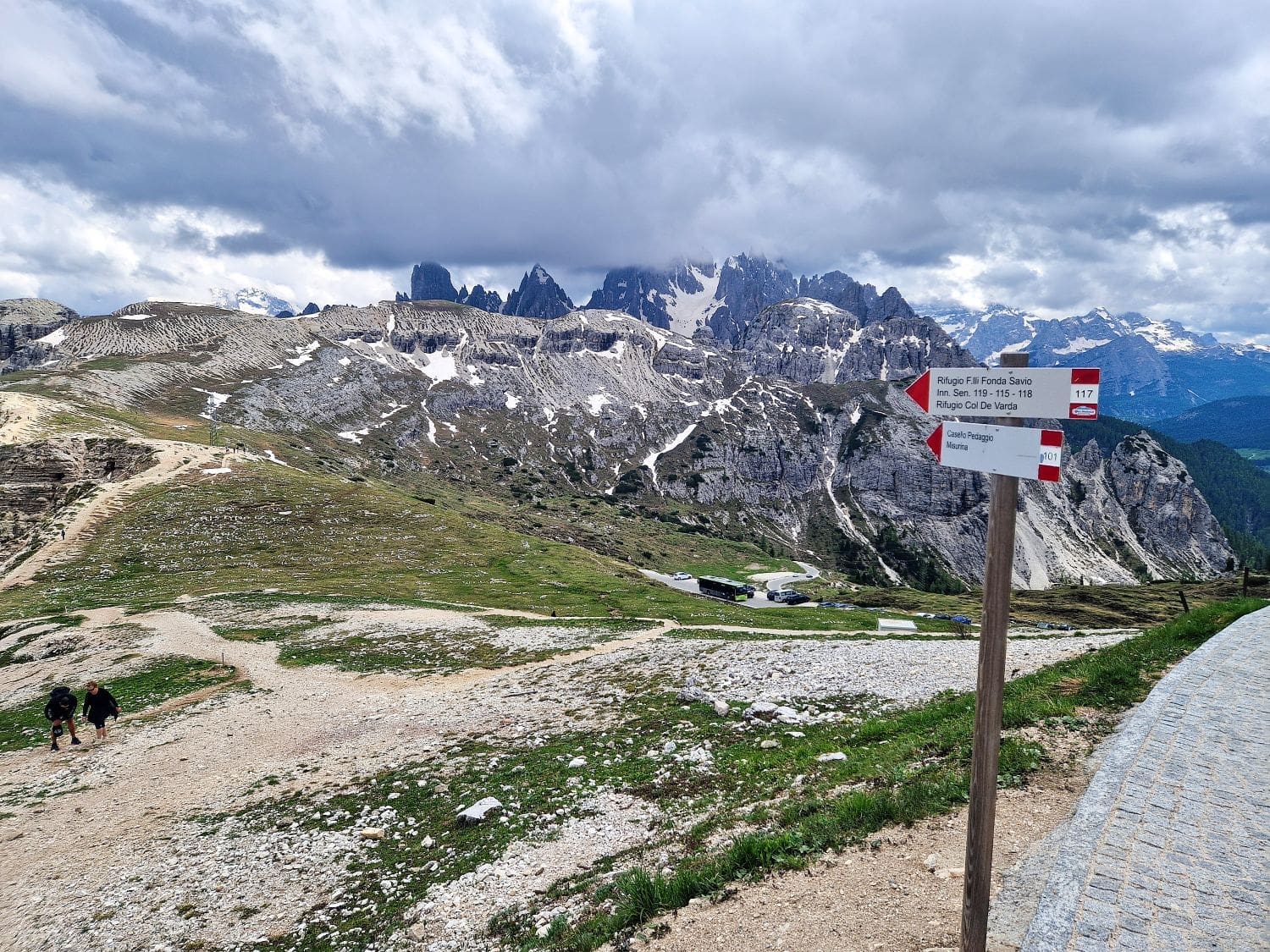 A mountainous landscape with dramatic cloud cover. Rugged, snow-capped peaks dominate the background, including the renowned Cadini di Misurina viewpoint. In the foreground, a signpost shows hiking directions. Two hikers walk along a rocky path to the left, while a winding paved trail leads to the right.