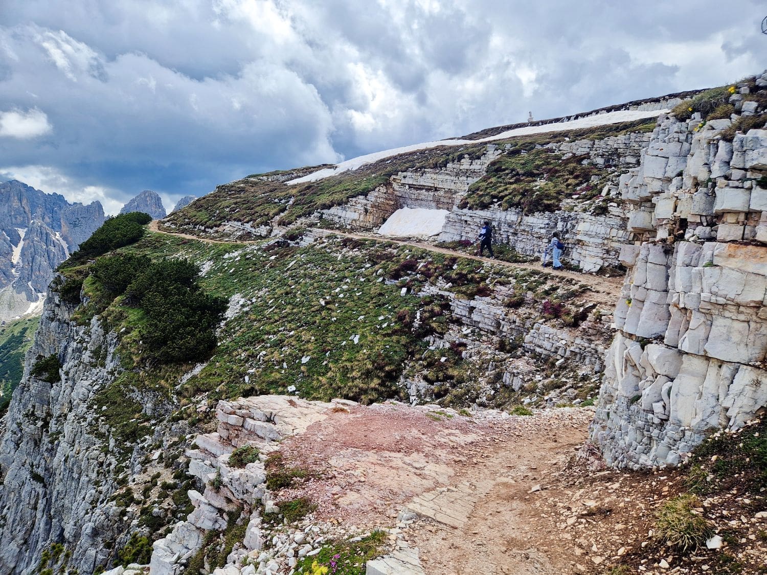 A narrow, rocky mountain trail winds along a steep cliffside with patches of greenery and snow. Two hikers are present on the path, likely taking in the breathtaking views offered by the Cadini di Misurina Viewpoint hike. In the distance, jagged mountain peaks are seen under a cloudy sky.