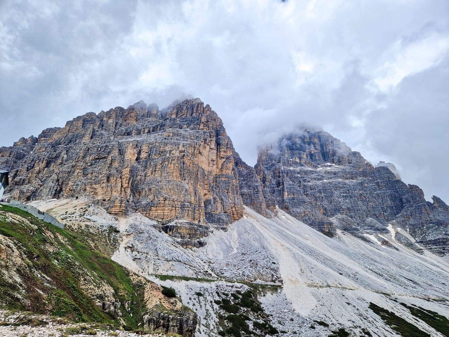 A rugged mountain range with steep, rocky cliffs partially covered by mist and clouds. The foreground features stony slopes with patches of grass and small shrubs. From the Cadini di Misurina viewpoint hike, the overcast sky adds a dramatic effect to the scene.