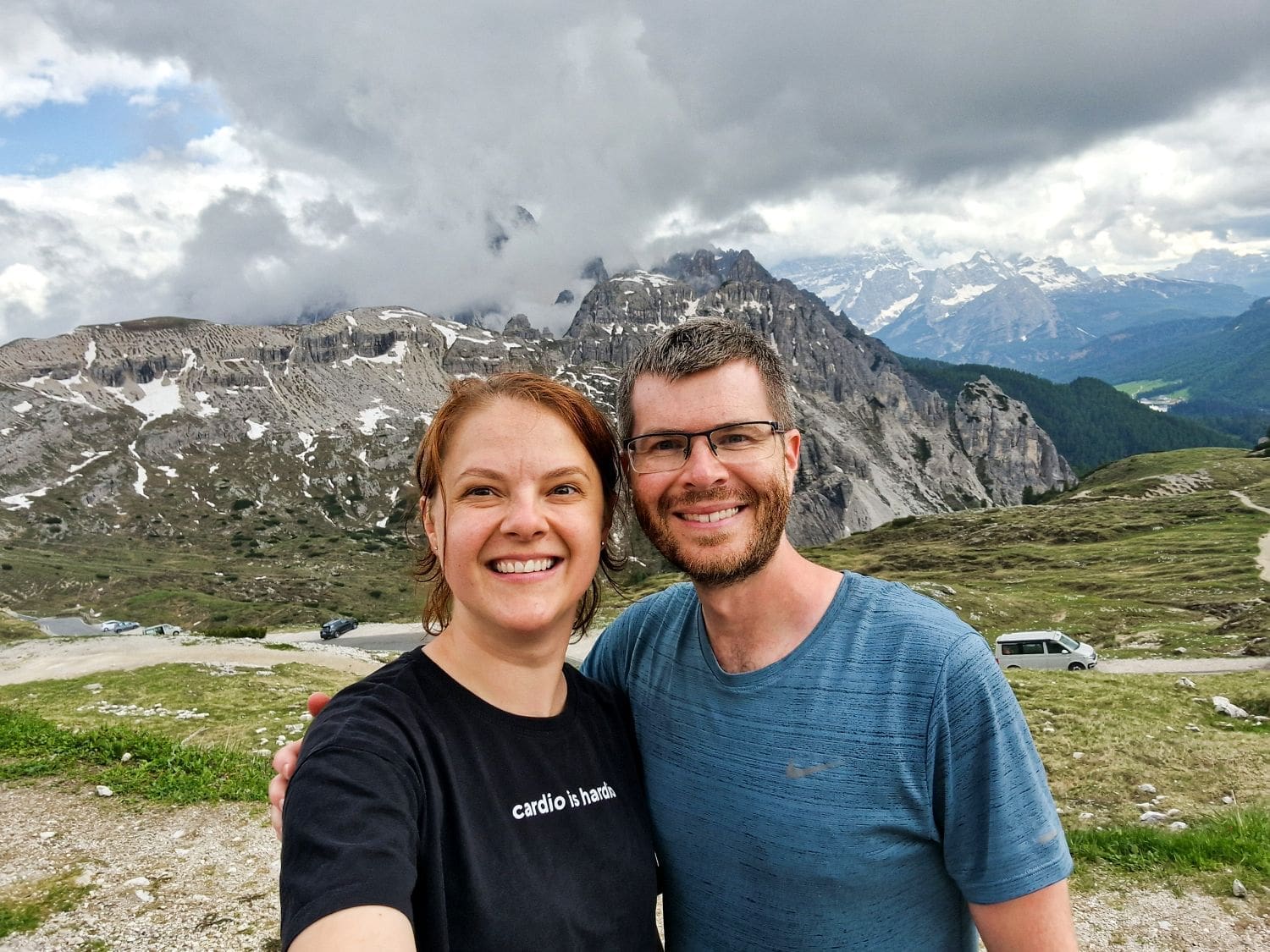 A smiling couple stands close together, taking a selfie in front of a picturesque mountain landscape. Snow-capped peaks, green valleys, and a cloudy sky form the stunning backdrop. The man wears glasses and a blue shirt, while the woman wears a black t-shirt.