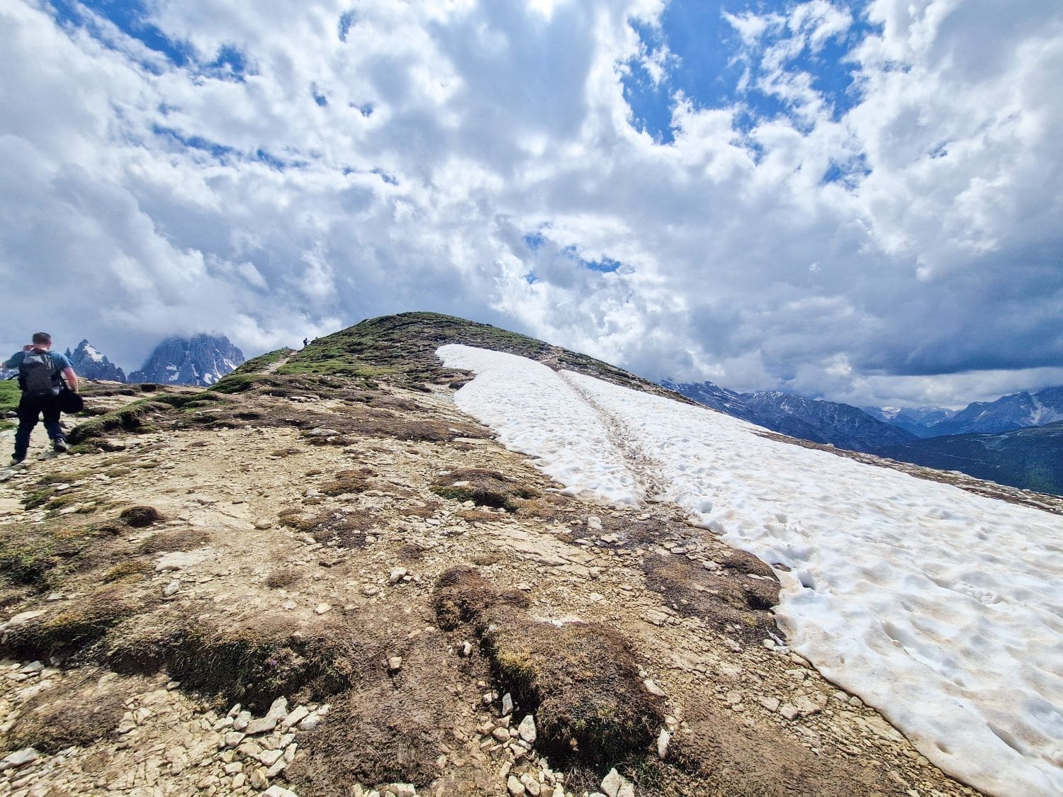 A hiker walks up a rocky path toward a snow-covered section of a mountain under a cloudy sky. Snow patches contrast with the bare ground, and distant peaks are visible under the dramatic clouds, capturing the breathtaking essence of the Cadini di Misurina viewpoint hike.