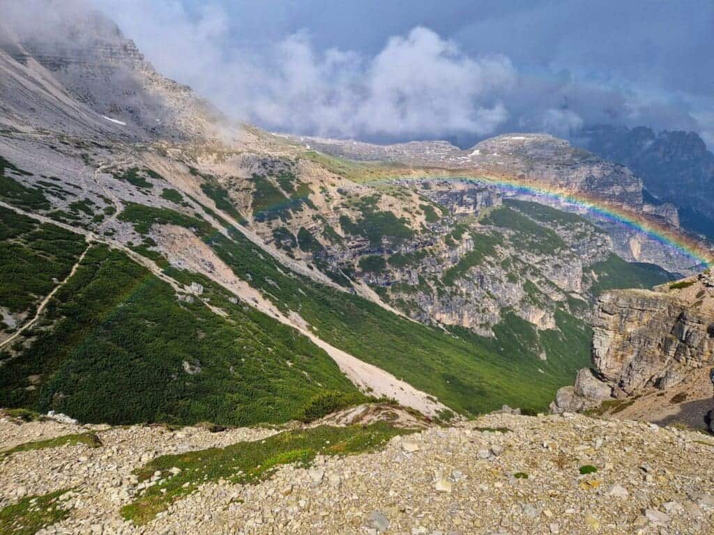 A stunning mountain landscape with rocky terrain and patches of green vegetation is shown under a cloudy sky at the Cadini di Misurina Viewpoint. A vibrant rainbow arcs across the scene, intersecting the cliffs on the right and fading away into the mist on the left.