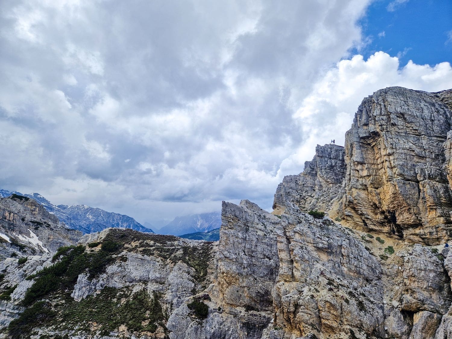 A scenic view of rugged, steep cliff faces under a partly cloudy sky. Small figures of people are visible standing atop one of the cliffs at the Cadini di Misurina viewpoint, highlighting the scale and grandeur of the landscape. The distant mountains and patches of greenery add depth to the scene.