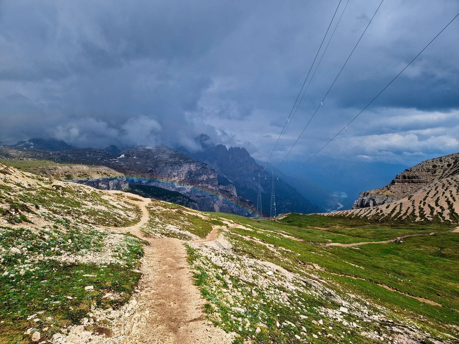 A rocky mountain trail winds through green grass under a stormy, cloud-filled sky. Power lines run along the right side of the image. In the distance, mist shrouds the Cadini di Misurina viewpoint peaks, and a faint rainbow can be seen.