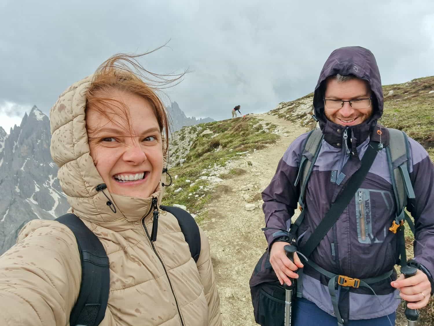 Two people dressed in outdoor clothing, including jackets and backpacks, smile and pose on a narrow mountain trail. The woman on the left has windblown hair, and the man on the right holds trekking poles. Rugged, cloudy mountain scenery is visible in the background at Cadini di Misurina Viewpoint.