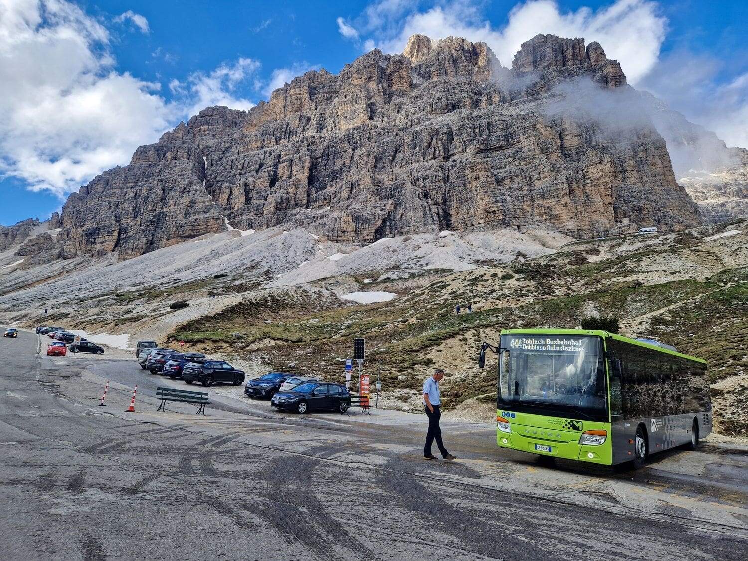 Are the Dolomites worth visiting? Yes! Why? 15 A green tourist bus is parked on a mountain road near several other cars, likely at the Cadini di Misurina viewpoint. A person is walking beside the bus. The rocky mountain landscape in the background is partially covered by clouds, with a clear blue sky above.