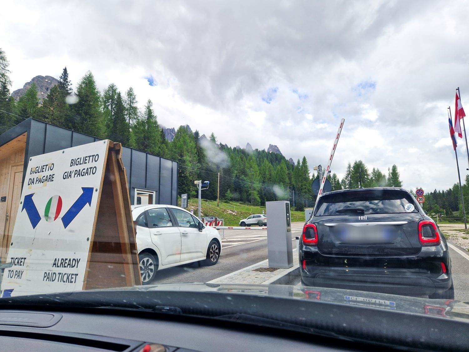 Cars are lined up at a toll booth in a mountain setting with trees and cloudy sky, reminiscent of the scenery you'd encounter on a cadini di misurina viewpoint hike. A sign in Italian and English directs drivers to pay tickets or pass with pre-paid tickets. Flags are visible, and a barrier arm is raised to let a car through.