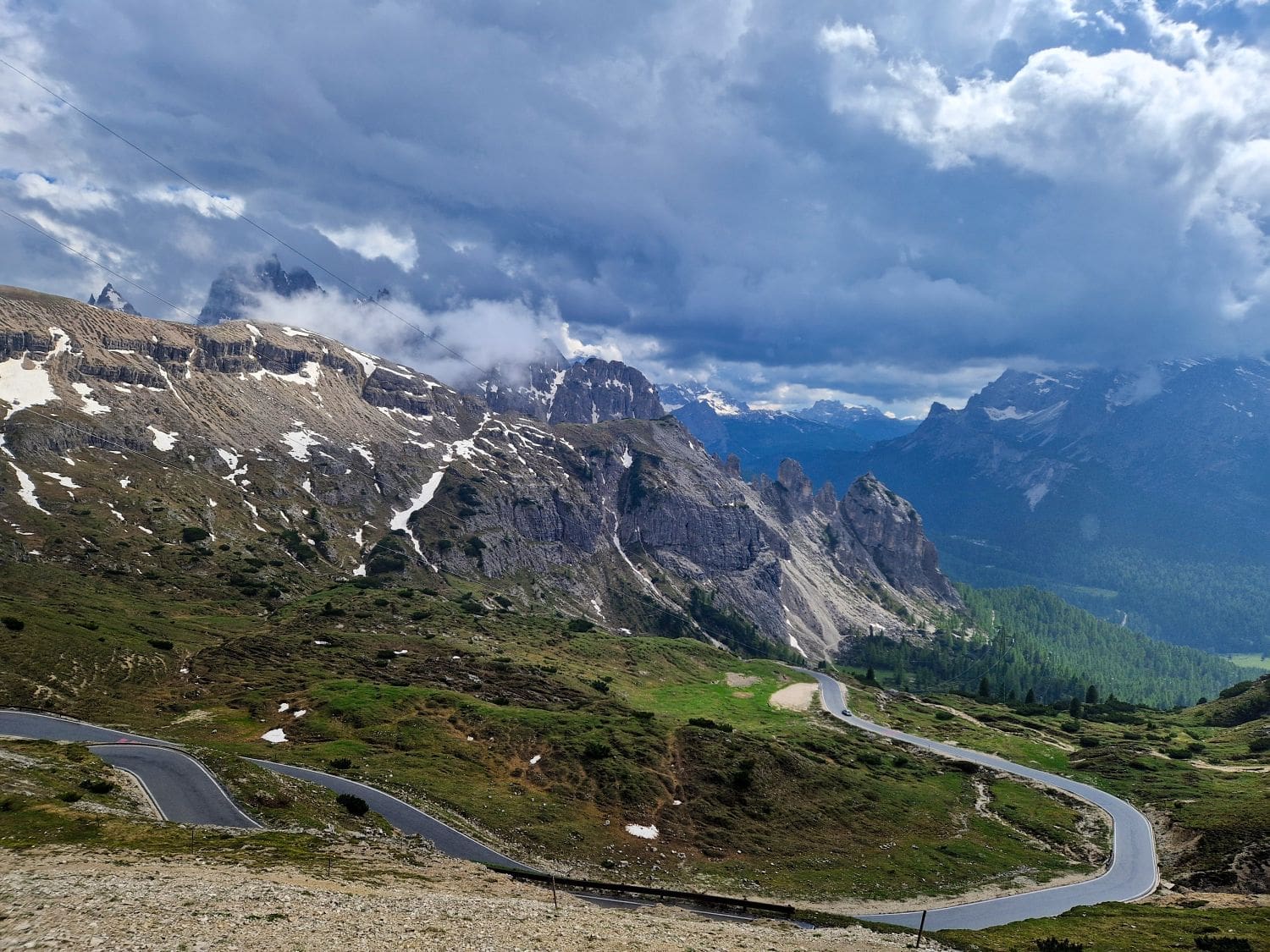 A picturesque mountainous landscape unfolds at the Cadini di Misurina viewpoint, where a winding road cuts through green meadows. Snow-capped peaks and rocky cliffs rise under a cloudy sky, with patches of greenery and distant lush forests adding to the stunning backdrop.