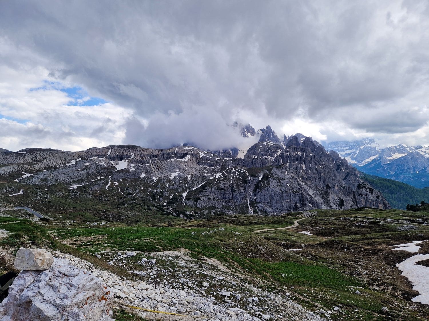 A breathtaking view of the mountainous landscape at the Cadini di Misurina, with rocky peaks partially covered by clouds. The lower slopes show patches of snow and green vegetation. A meandering path winds through the terrain, adding depth and leading the eye towards the distant mountains.