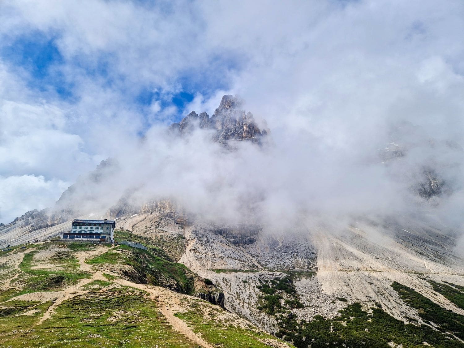 A serene mountainous landscape features rugged peaks partially shrouded in clouds, reminiscent of the views from the Cadini di Misurina viewpoint. A small, isolated building sits prominently on a grassy slope, while rocky terrain and patches of vegetation complete the scene under a partly cloudy blue sky.