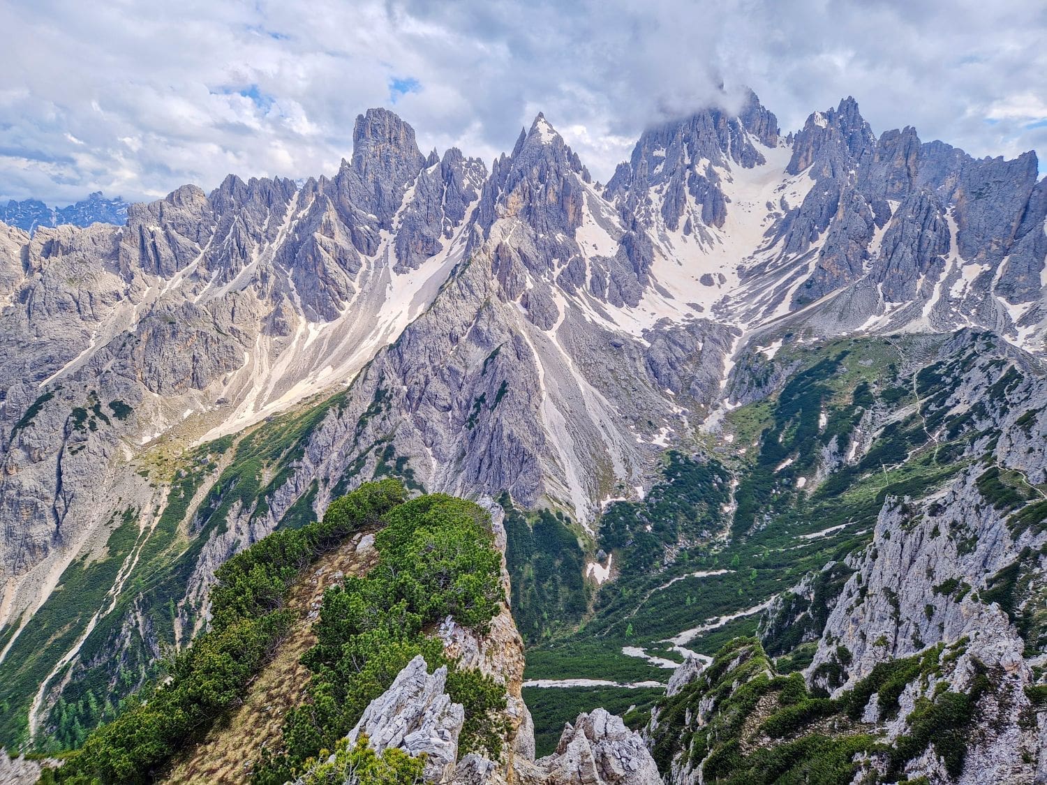 A breathtaking view of the mountain range from the Cadini di Misurina viewpoint, featuring jagged, snow-capped peaks under a partly cloudy sky. The foreground shows a rugged mountainside with patches of green vegetation, leading down to a lush, forested valley contrasted with rocky terrain.