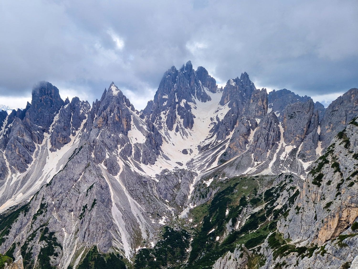 A rugged mountain range with sharp, jagged peaks covered in snow and patches of green vegetation, set against a cloudy sky.