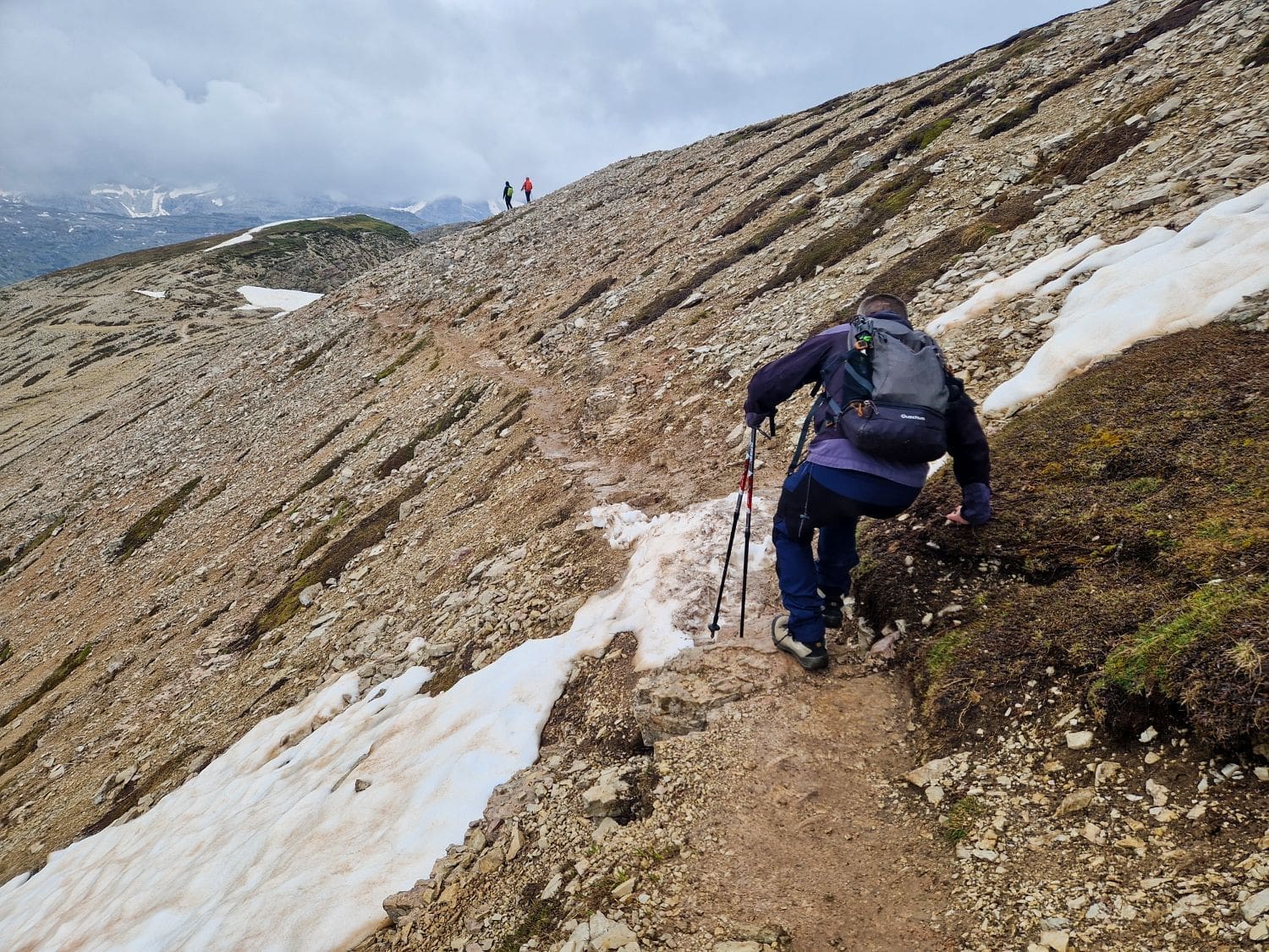 A hiker with a backpack and trekking poles ascends a rocky and snowy mountain trail. The terrain has patches of snow and loose rocks. Two other hikers are visible in the distance against a cloudy sky, walking towards the peak.