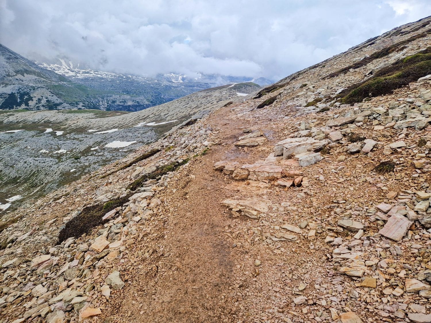 A mountain trail made of loose rocks and dirt winds through a rocky landscape with patches of snow. The overcast sky and scattered clouds suggest cool weather. Jagged peaks and valleys can be seen in the background, offering glimpses reminiscent of the stunning Cadini di Misurina viewpoint hike.