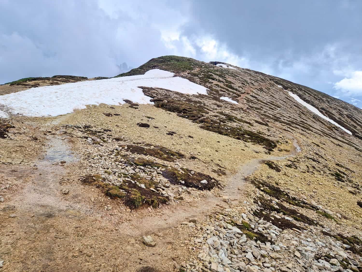 A narrow, winding trail leads up a rugged, rocky mountain slope. Patches of snow are scattered along the trail, contrasting with the earthy terrain. The sky above is overcast with gray clouds, suggesting an approaching storm—a perfect setting for a Cadini di Misurina viewpoint hike.