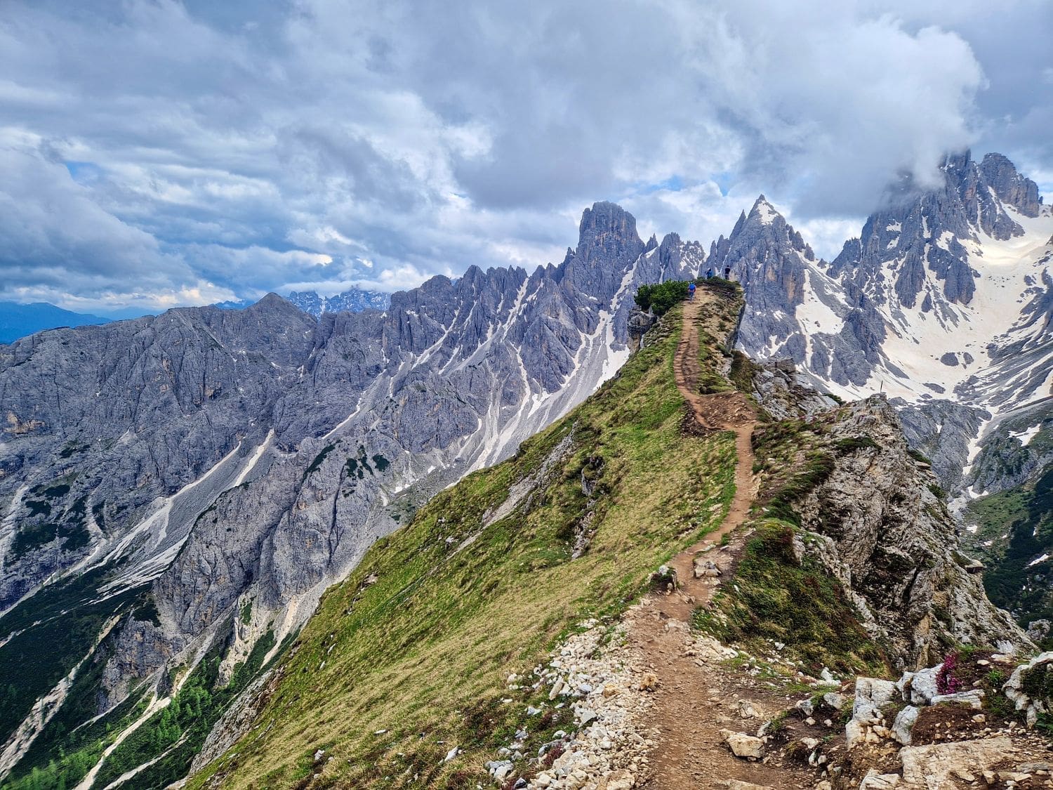 A rugged mountain path winds along a narrow ridge, surrounded by towering peaks partially covered in snow. The sky is overcast with thick clouds. Vegetation is sparse, and the rugged terrain is marked by rocky outcrops, typical of the renowned Cadini di Misurina viewpoint hike.