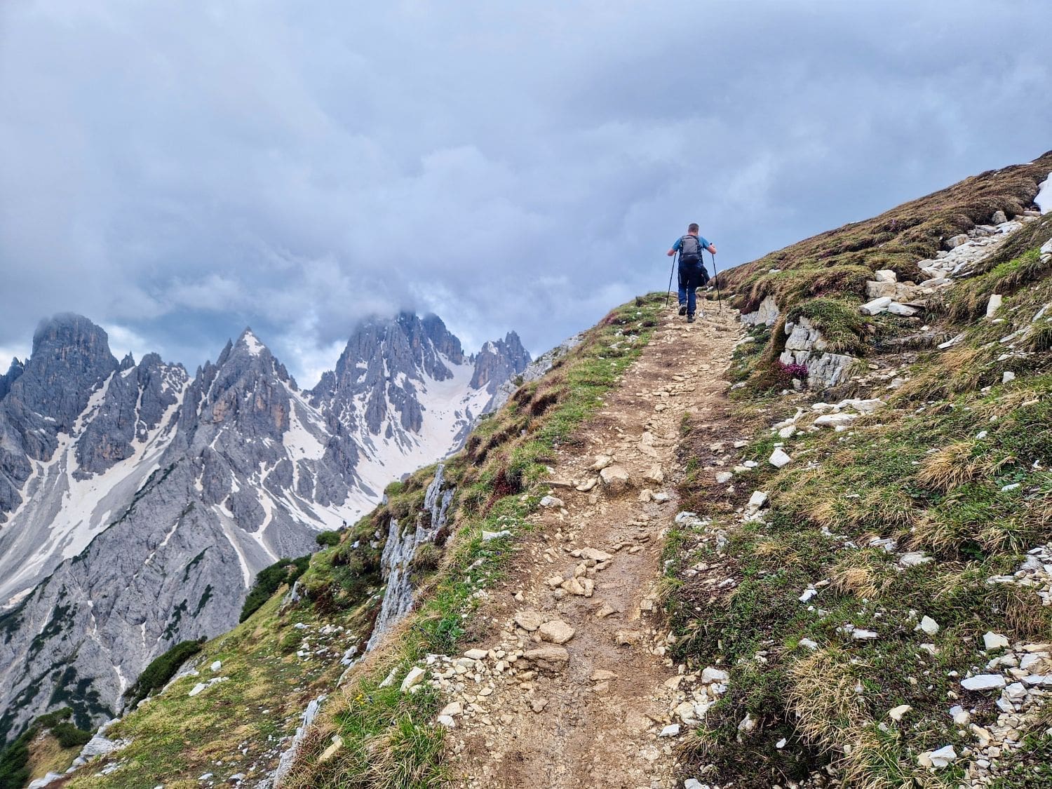 A hiker with a backpack and trekking poles ascends a rocky mountain trail. Snow-capped peaks and dramatic, jagged mountains stand under a cloudy sky in the background. Sparse greenery and grass patches appear along the path, leading towards the stunning Cadini di Misurina viewpoint hike.