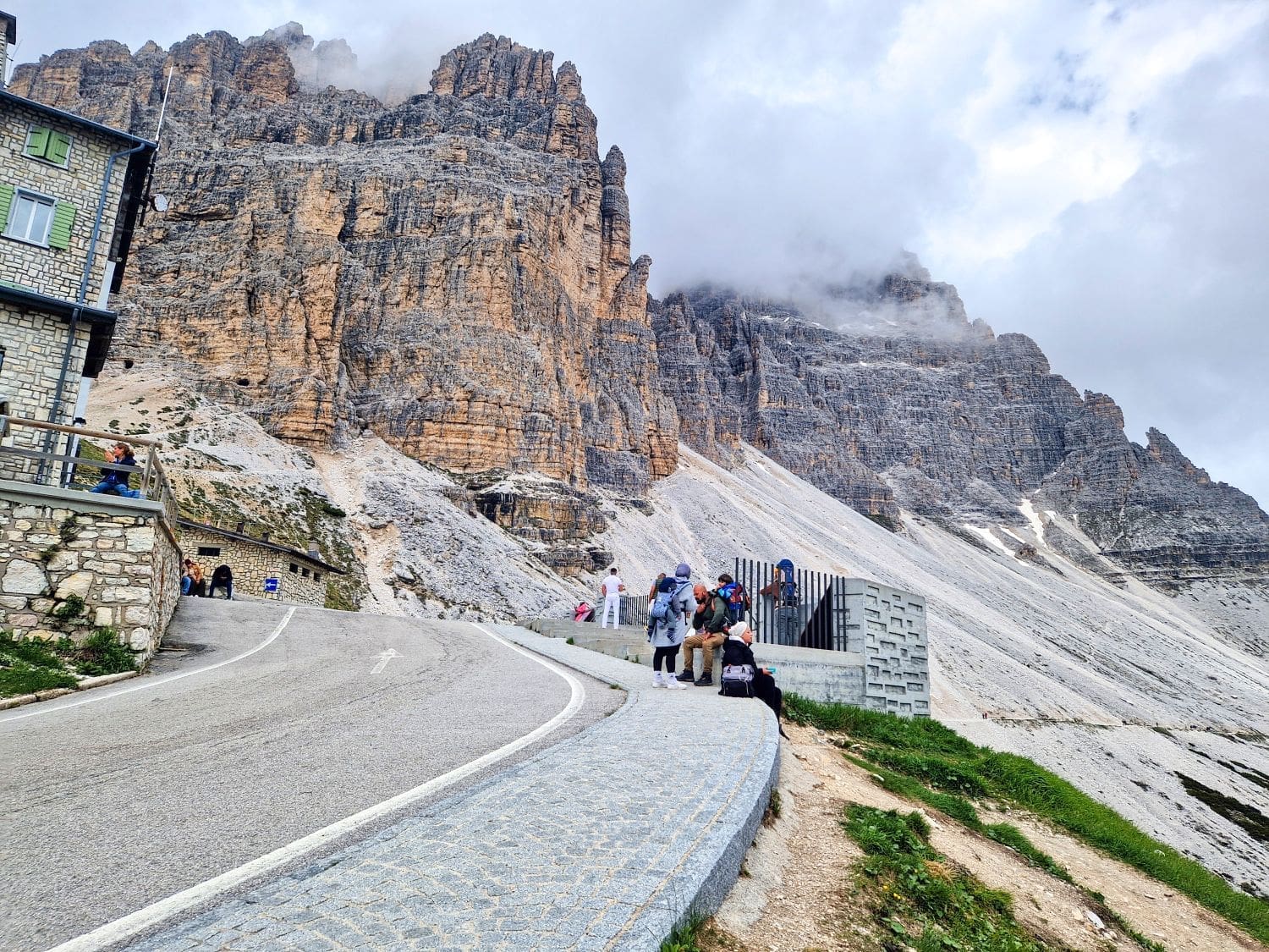 A scenic mountainous landscape featuring a winding road leading to the Cadini di Misurina viewpoint hike, where several people are gathered. Towering rocky cliffs dominate the background, partially obscured by clouds. A building sits on the left side of the road near the cliffs.