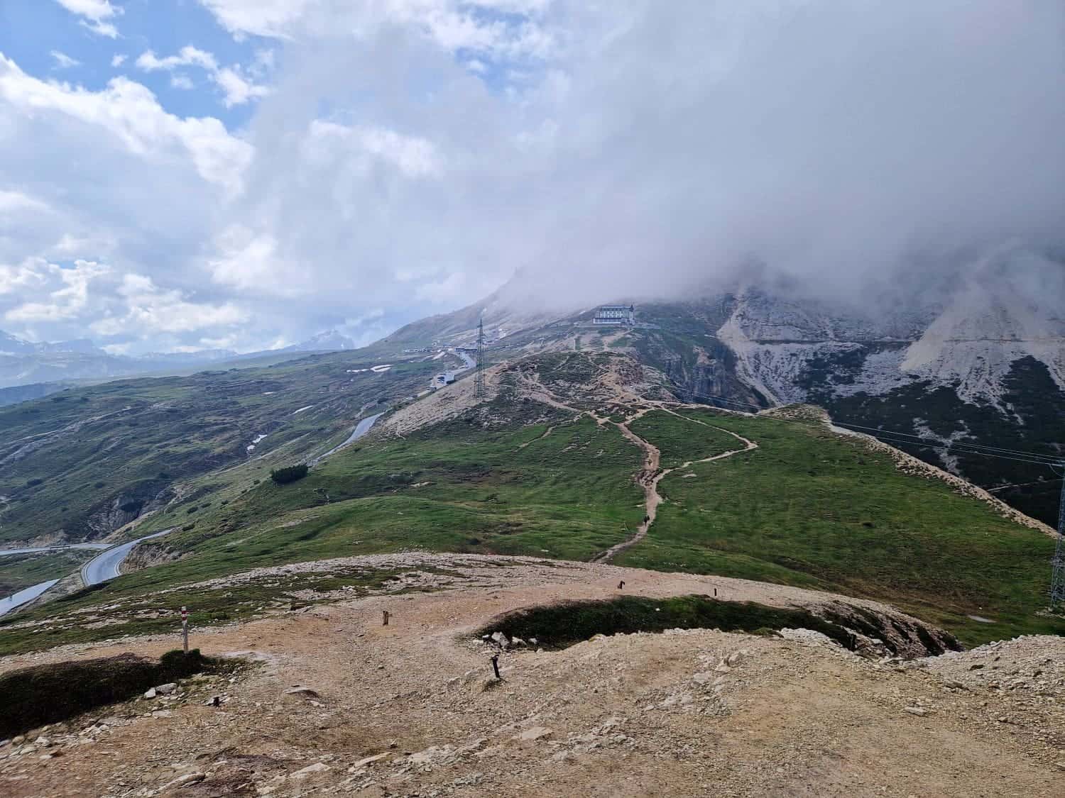 A scenic mountainous landscape with winding trails and pathways leading up the hills. The sky is partly cloudy, with some low-lying clouds obscuring the peaks. Green fields and rocky terrain can be seen in the foreground. Tiny figures of hikers heading towards the Cadini di Misurina viewpoint are visible.
