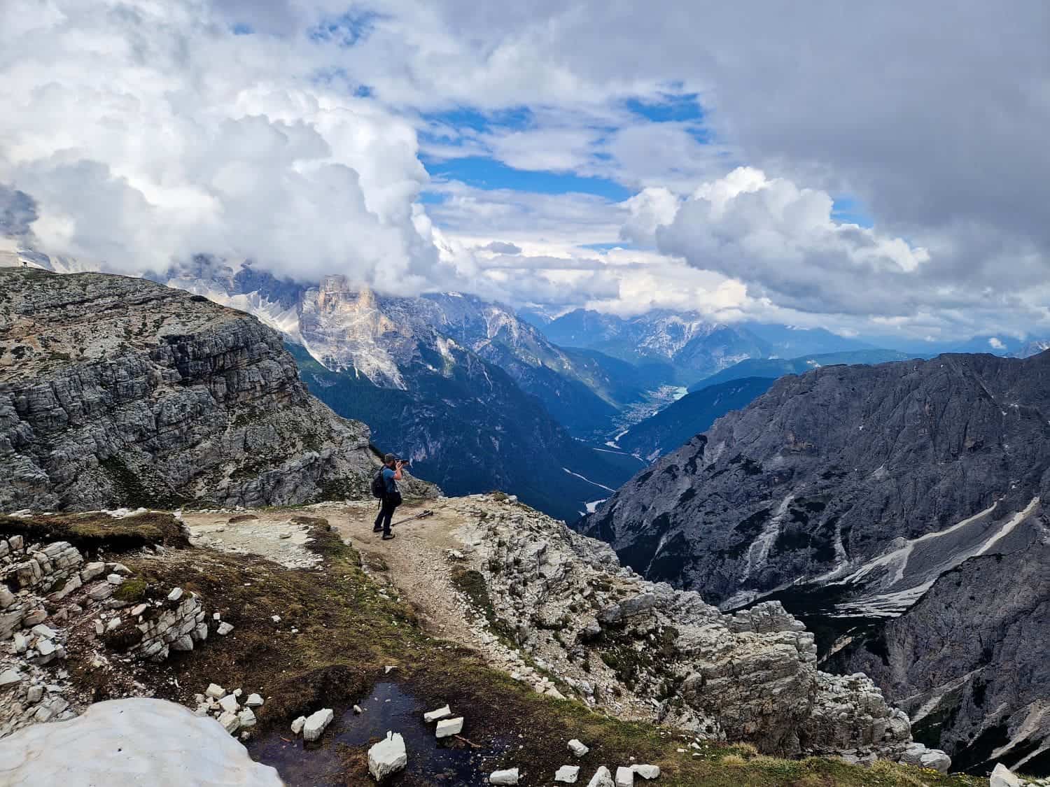 A hiker stands on a rocky, narrow mountain trail at the Cadini di Misurina viewpoint, surrounded by jagged peaks and cliffs. The sky above is partly cloudy, revealing patches of blue. In the distance, lush valleys and distant mountains are visible. The scene is one of rugged, natural beauty.