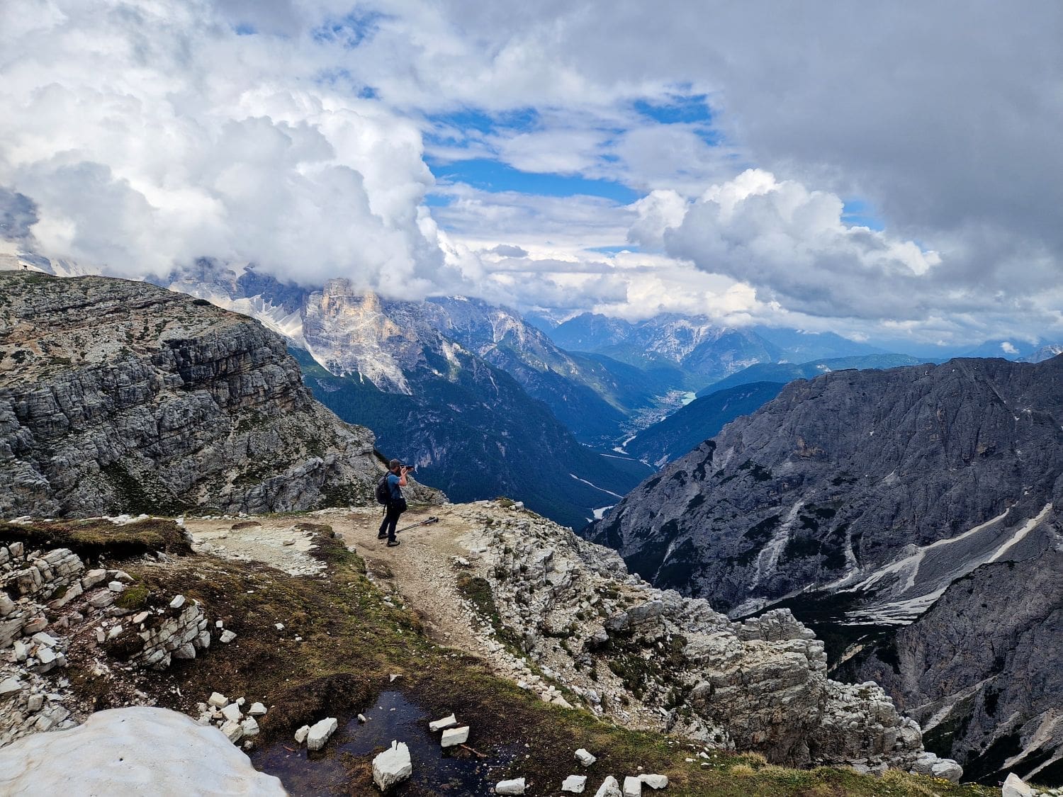 A hiker stands on a rocky, narrow mountain trail at the Cadini di Misurina viewpoint, surrounded by jagged peaks and cliffs. The sky above is partly cloudy, revealing patches of blue. In the distance, lush valleys and distant mountains are visible. The scene is one of rugged, natural beauty.