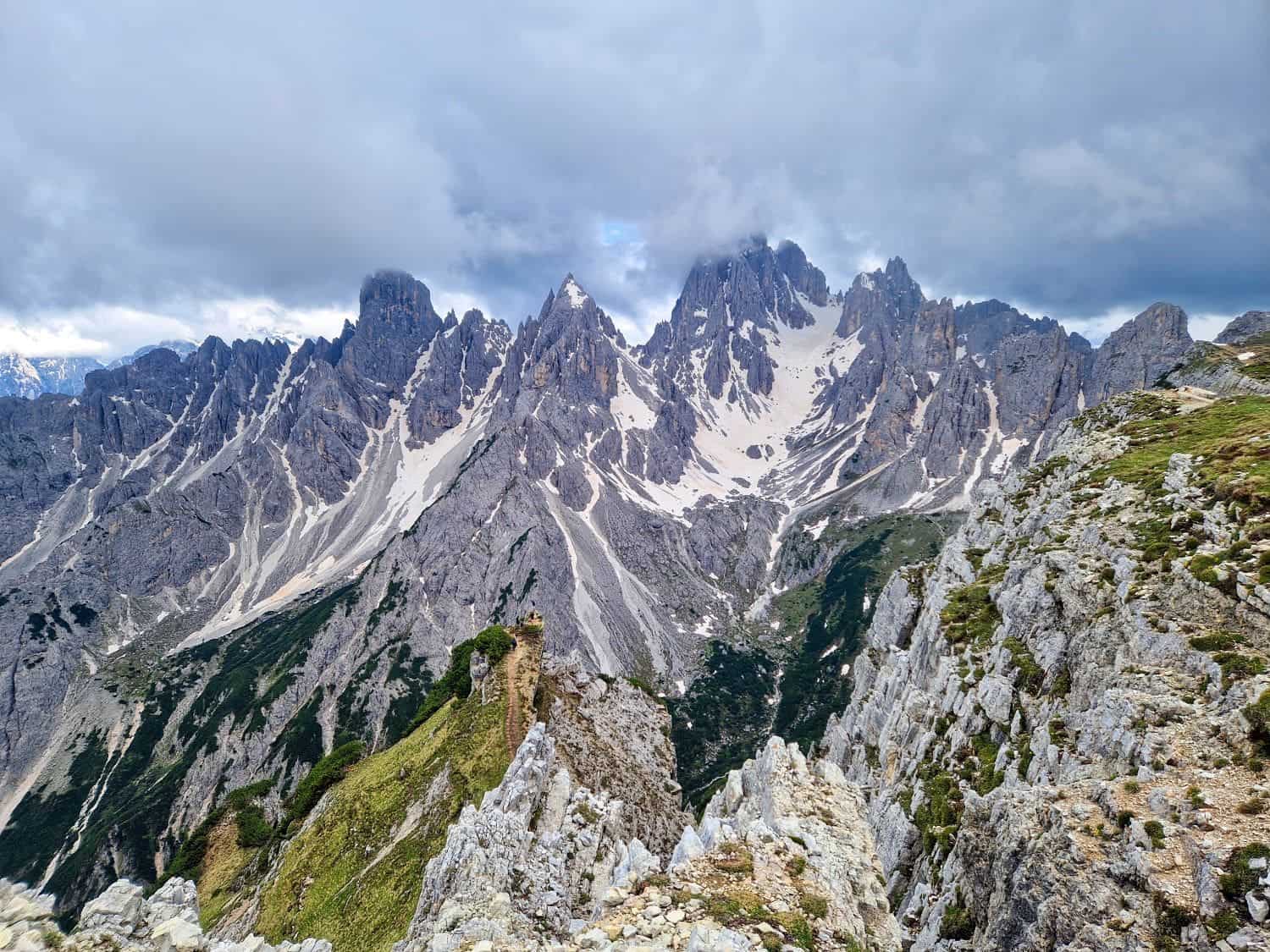 A rugged mountain range with jagged, snow-capped peaks under a cloudy sky. The Cadini di Misurina viewpoint hike reveals sharp cliffs and ridges amid patches of green vegetation. In the foreground, a rocky trail winds through the terrain, offering breathtaking vistas at every turn.
