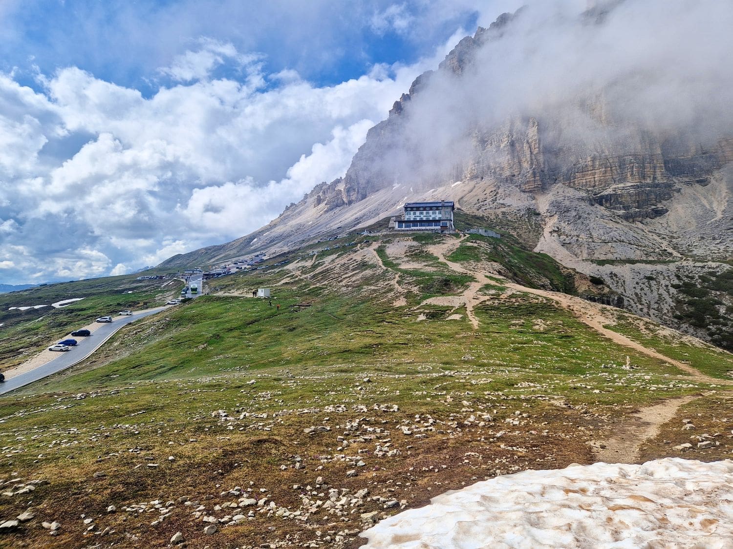 A scenic mountain landscape with a winding road leading to a building nestled against rocky cliffs. Clouds partially cover the mountain peaks, while patches of green grass and rocky terrain dominate the foreground. A few cars are visible on the road, offering a glimpse of the Cadini di Misurina viewpoint hike nearby.