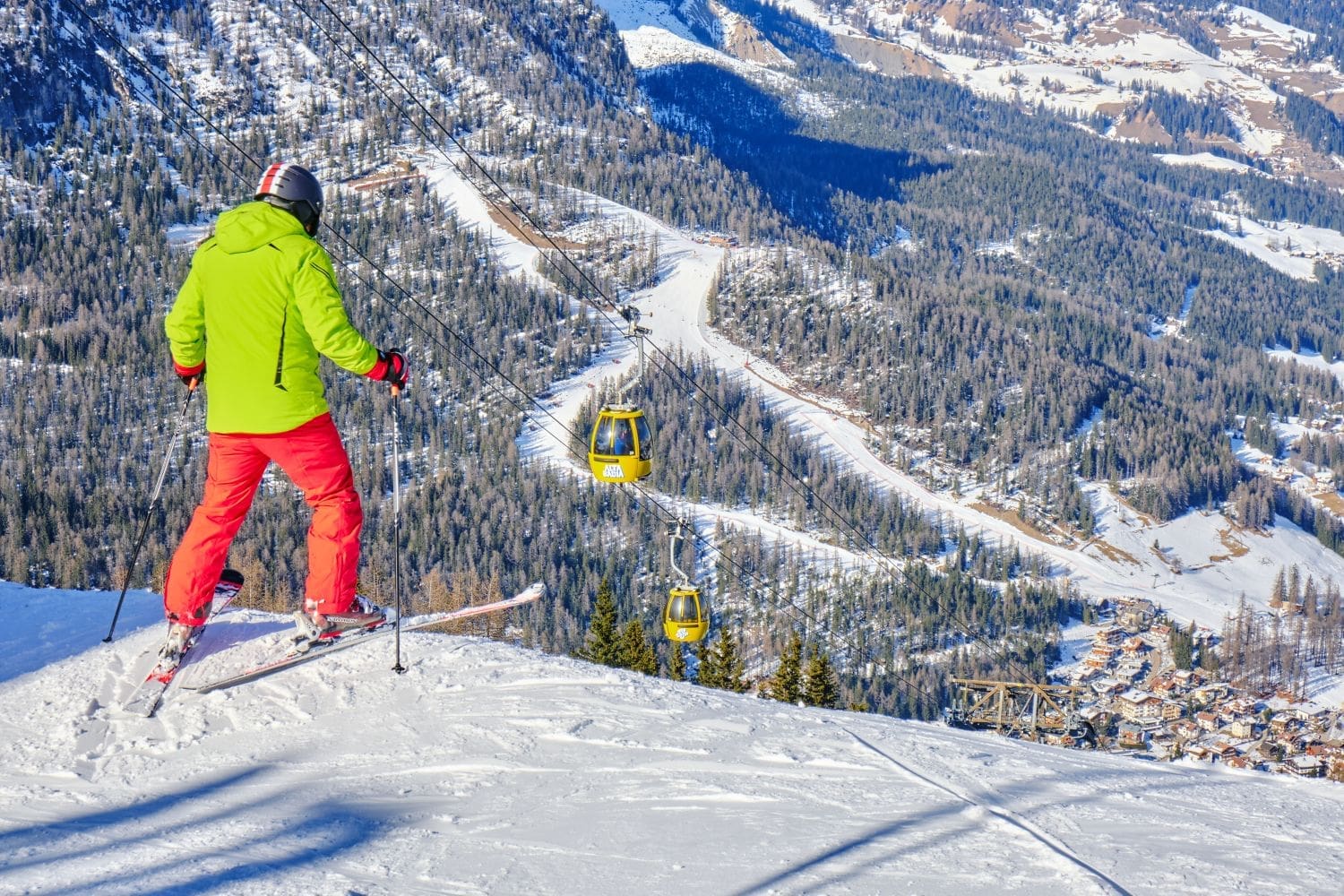 A skier in a green jacket and red pants looks down a snowy slope with yellow gondolas suspended on cables in front. Snow-covered trees and a mountain village are visible in the background under a clear blue sky.