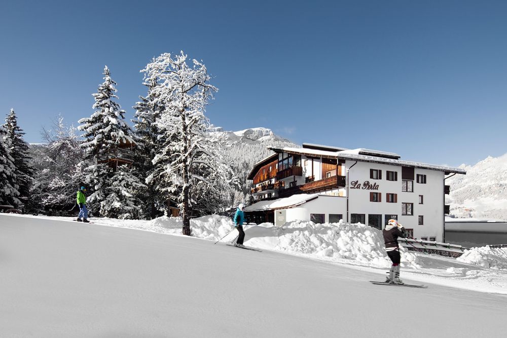 Three people ski on a snowy slope near a chalet-style hotel surrounded by snow-covered trees and mountains under a clear blue sky.