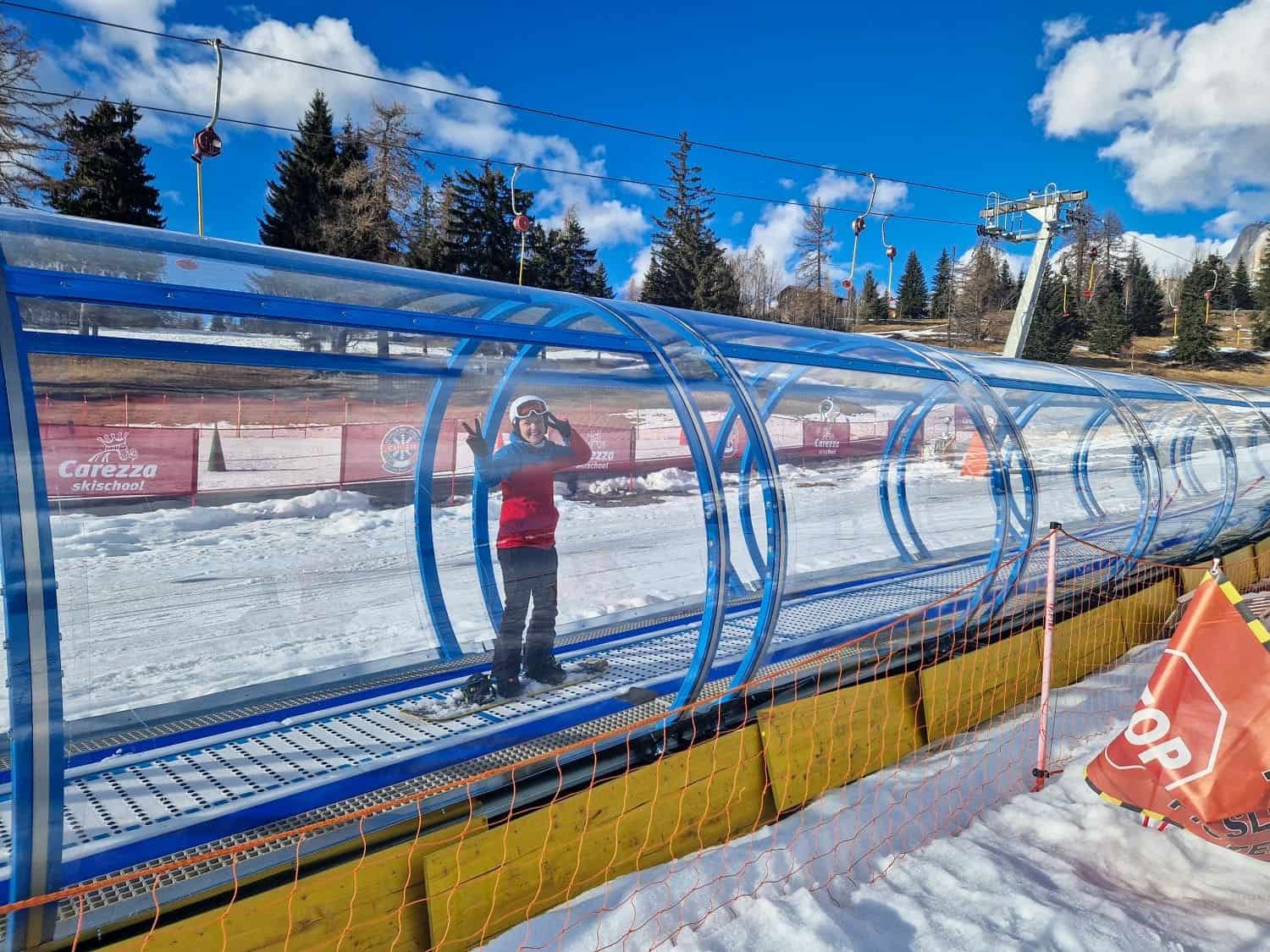 A person in a red jacket is standing on a covered conveyor belt ski lift in a snowy area. The sky is clear and blue with a few scattered clouds. Orange safety netting is visible in the foreground, and there are pine trees and ski equipment in the background.