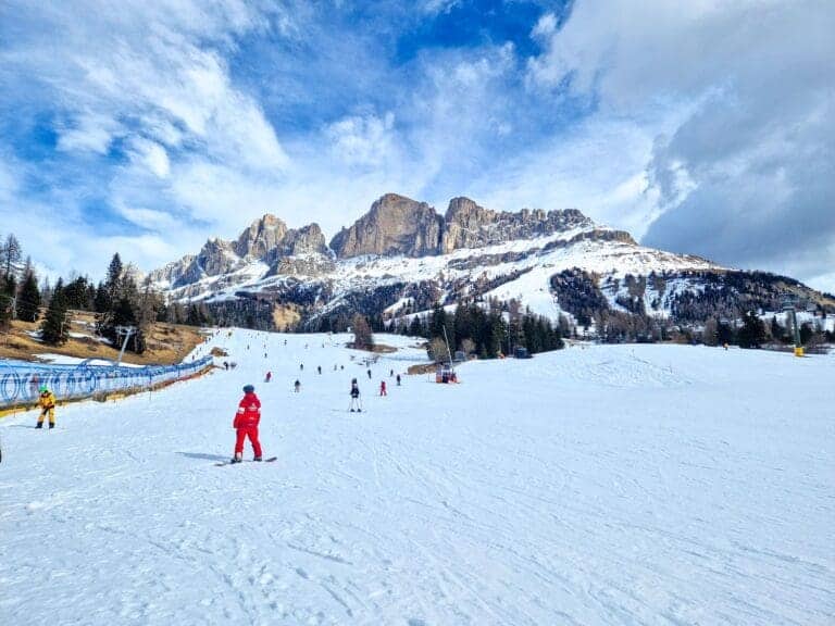 A snow-covered ski slope with skiers and snowboarders, surrounded by mountainous terrain. In the foreground, a person in a red jacket is skiing. Pine trees line the sides of the slope, and a dramatic sky with clouds and patches of blue is overhead.