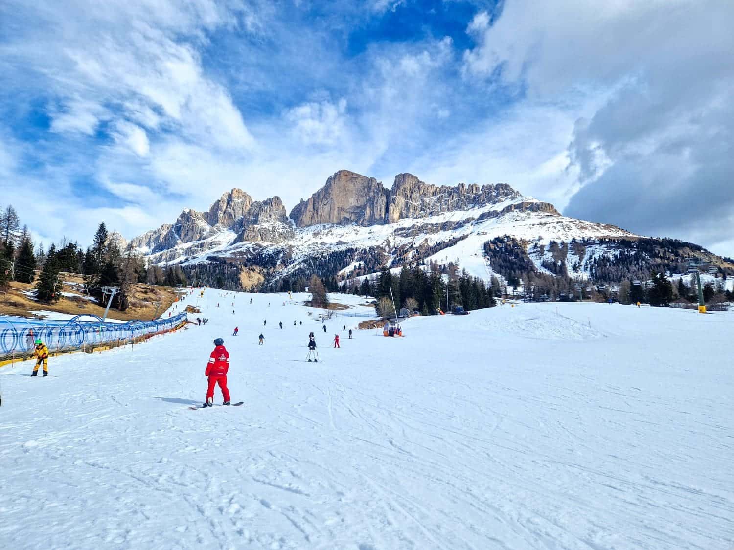 A snow-covered ski slope with skiers and snowboarders, surrounded by mountainous terrain. In the foreground, a person in a red jacket is skiing. Pine trees line the sides of the slope, and a dramatic sky with clouds and patches of blue is overhead.
