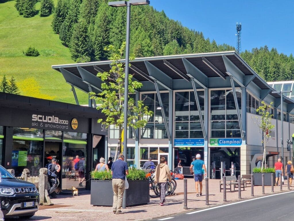 A modern building serves as the lift station for the Ciampinoi cable car in a mountainous area. People walk and ride bikes in front of the station, while sunshine illuminates nearby shops, including "Scuola 2000." Lush green hills and pine trees form the scenic backdrop.