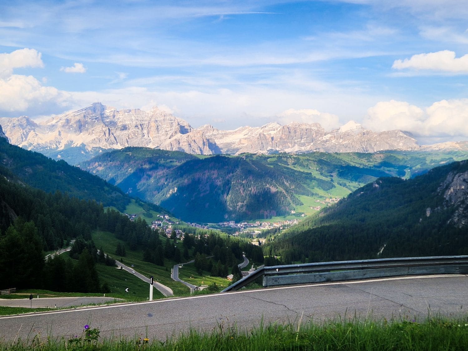 A scenic view of winding roads leading through a lush, green valley surrounded by forested hills and distant snow-capped mountains under a blue sky with scattered clouds. A metal guardrail lines the roadside in the foreground.