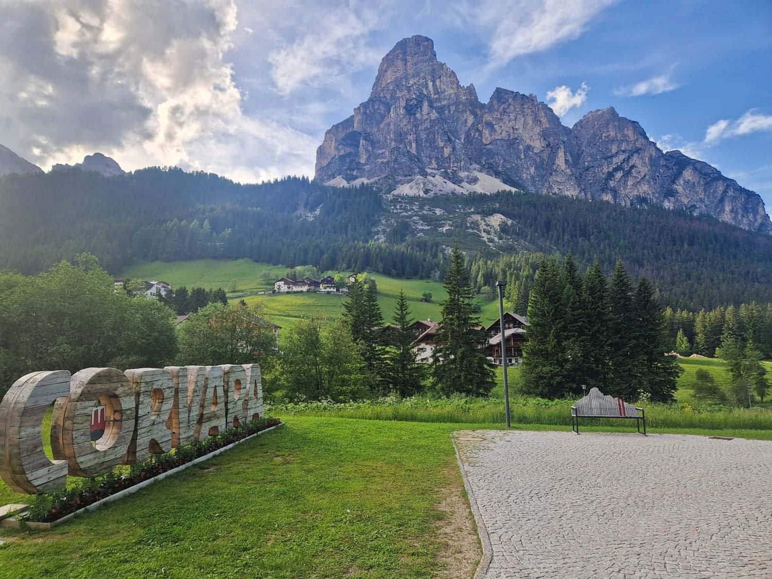 A picturesque scene of Corvara, Italy, featuring a large wooden sign reading "CORVARA" on the left, lush green grass, a cobblestone path, and a wooden bench. In the background, houses are nestled among dense forests with towering jagged mountains under a partly cloudy sky.
