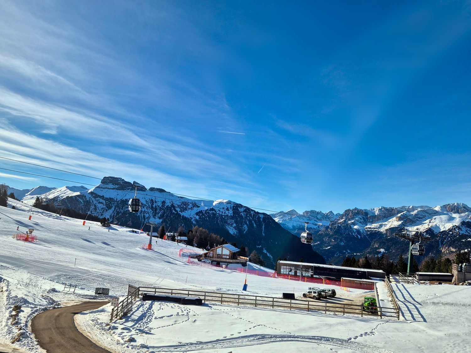 Wide view of a snowy ski resort with groomed slopes gondola cabins moving across the scene and jagged mountain peaks in the distance under a bright blue sky. A lodge and lift station sit near the base of the slope with fencing vehicles and ski tracks visible in the foreground. The image shows the scale of the landscape and the lift access that connects skiers to different areas of the mountain.