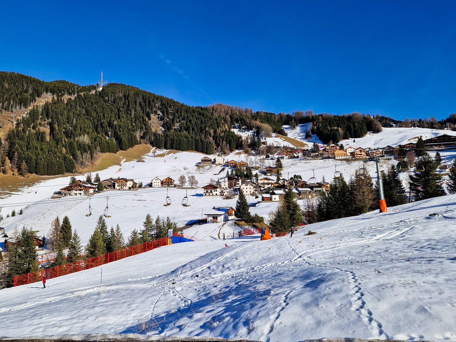 Snowy ski slope overlooks a small alpine village with clustered chalets scattered across the hillside and chairlifts running above the terrain. Evergreen forests line the upper slopes while groomed runs fencing and ski tracks are visible in the foreground. The image shows how the ski area flows directly into the mountain village below.
