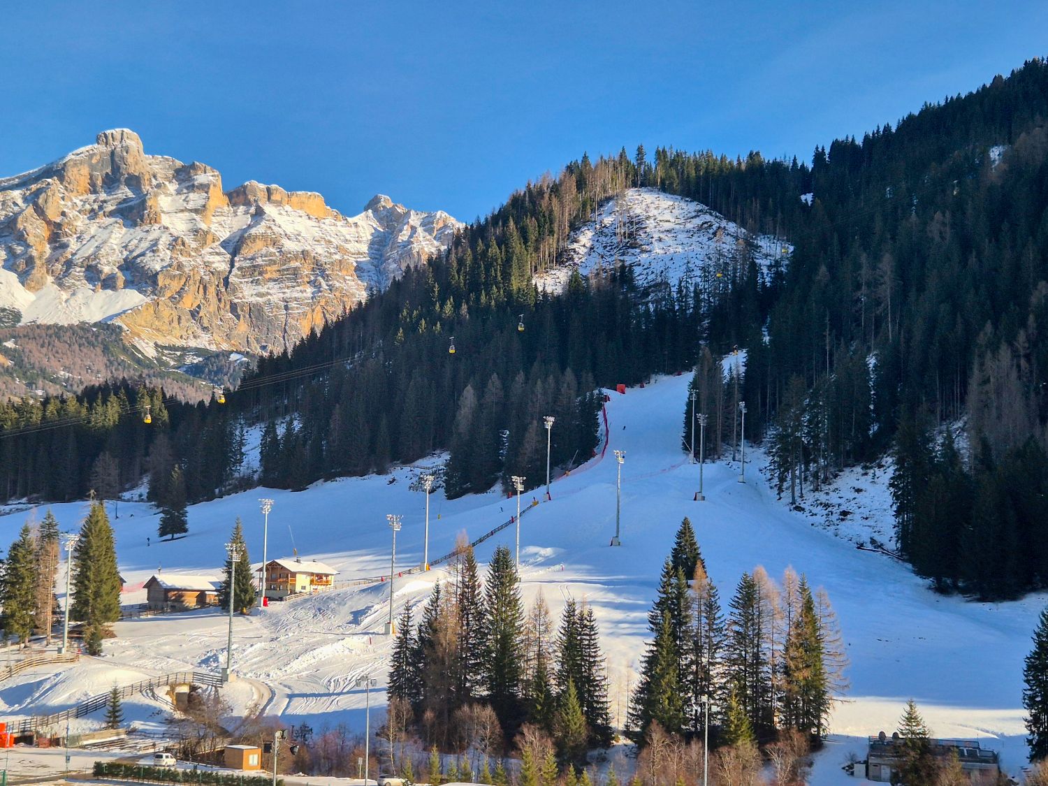 Snowy ski run cuts through a forested mountainside with tall evergreens lining the slope and lift towers rising from the valley below. A small ski lodge sits near the base while gondola cabins travel across the scene toward rugged rocky peaks in the background. The image highlights the contrast between groomed ski terrain dense forest and dramatic alpine cliffs.
