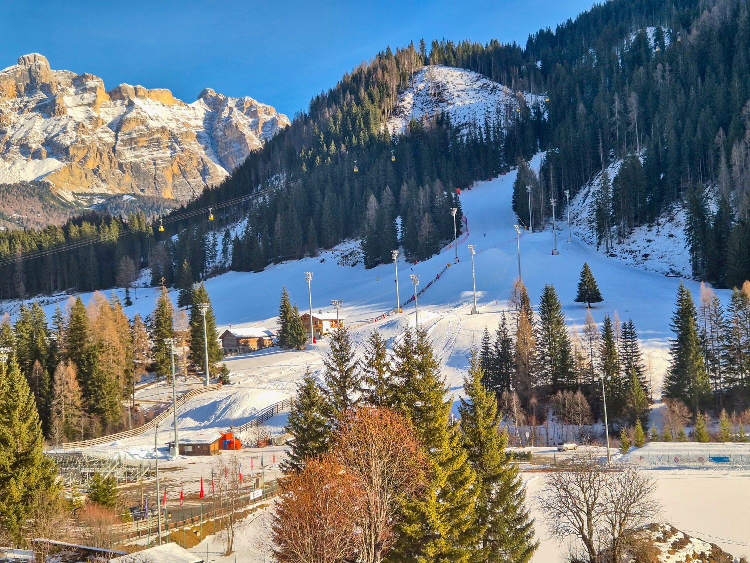 A sunlit ski area sits in a forested mountain valley with groomed slopes cutting through evergreen trees. Yellow gondola cars glide across the hillside while ski lifts buildings and light poles line the snow covered runs beneath rugged rocky peaks.
