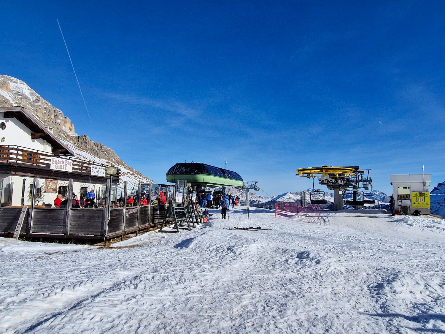 Snowy mountain ski area with a chairlift unloading zone and a round lift station at the top of the slope under a clear blue sky. On the left a busy mountain hut with an outdoor terrace is filled with skiers resting and socializing while skis are stacked nearby. The image shows a high altitude ski junction where lifts dining and trails meet.
