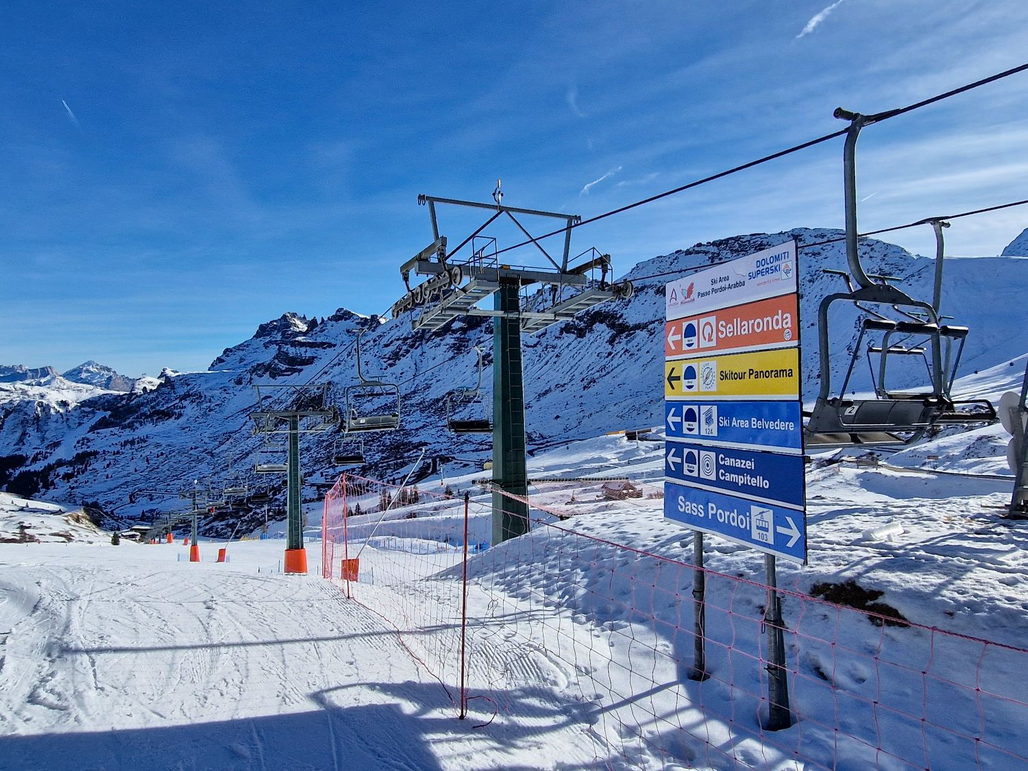 Snowy alpine ski area with a chairlift running overhead and rugged mountain peaks in the background under a clear blue sky. In the foreground a large directional sign reads Dolomiti Superski and shows arrows for Sellaronda, Skitour Panorama, Ski Area Belvedere, Canazei Campitello, and Sass Pordoi near Passo Pordoi Arabba. The image helps orient skiers by showing trail connections and lift access points at a mountain junction.