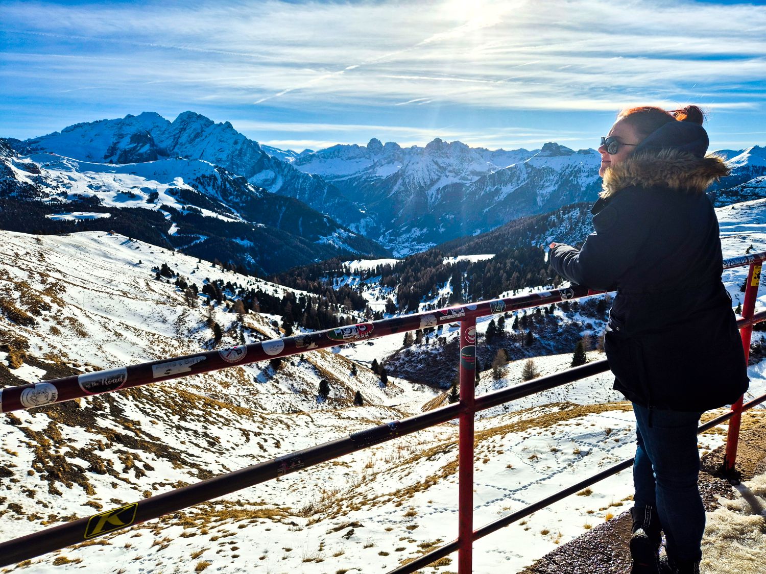 Person in a winter jacket stands at a mountain viewpoint looking out over a wide snow covered valley and jagged alpine peaks under a bright sky. A red metal railing lines the overlook while forested slopes and winding terrain drop away below. The image captures a quiet moment of scale and perspective in a high mountain landscape.