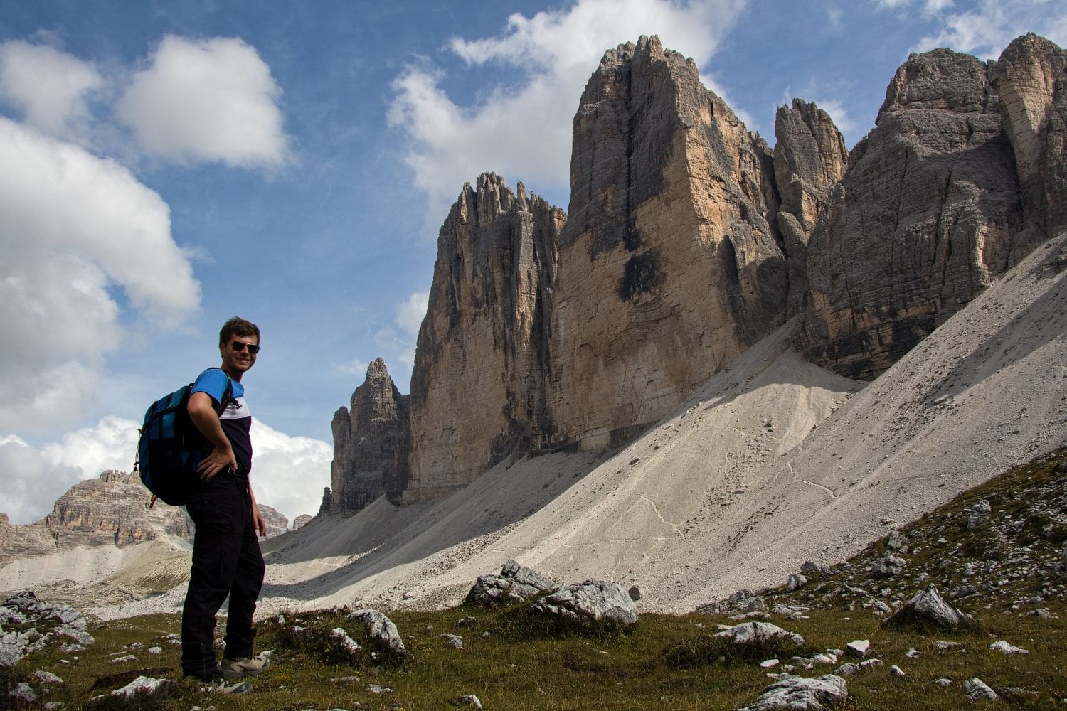 A hiker with a blue backpack stands on a grassy area, looking towards towering rock formations in the background under a partly cloudy sky. The scene suggests a rugged, mountainous landscape, possibly during a day hike or trek.