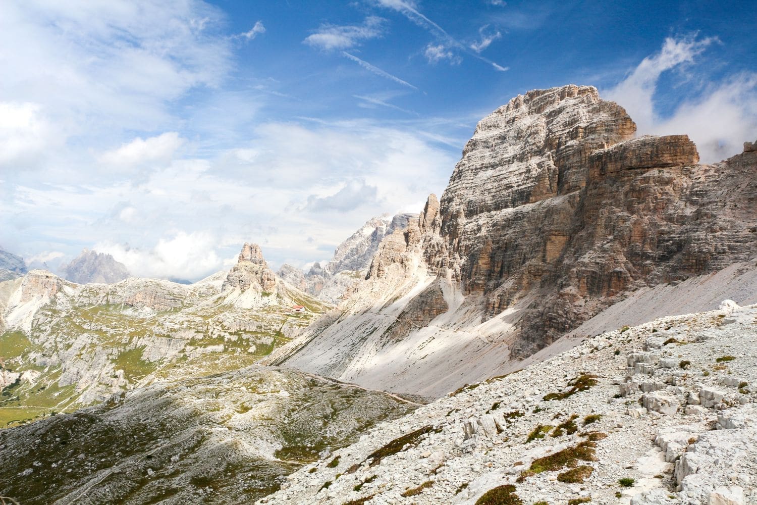 A mountainous landscape showing rugged, rocky peaks under a partly cloudy sky. The terrain is a mix of grey rocks and sparse green vegetation. A small red-roofed structure is visible in the distance, providing a sense of scale to the expansive view.