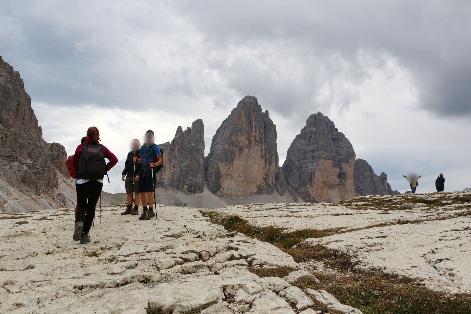 A group of hikers walking on a rocky trail towards three towering mountain peaks under a cloudy sky. One person in red jacket and hiking poles leads, followed by three others with backpacks. Another person stands in the distance, admiring the scenery.