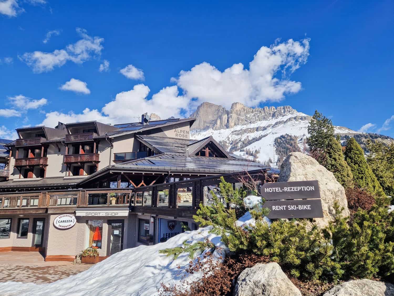 A cozy mountain lodge named Alpenrose nestled in the snowy Alps. The building has a rustic wood and stone exterior, with signs for hotel reception, ski and bike rental, and a pizzeria. Majestic snow-covered peaks rise in the background against a bright blue sky.