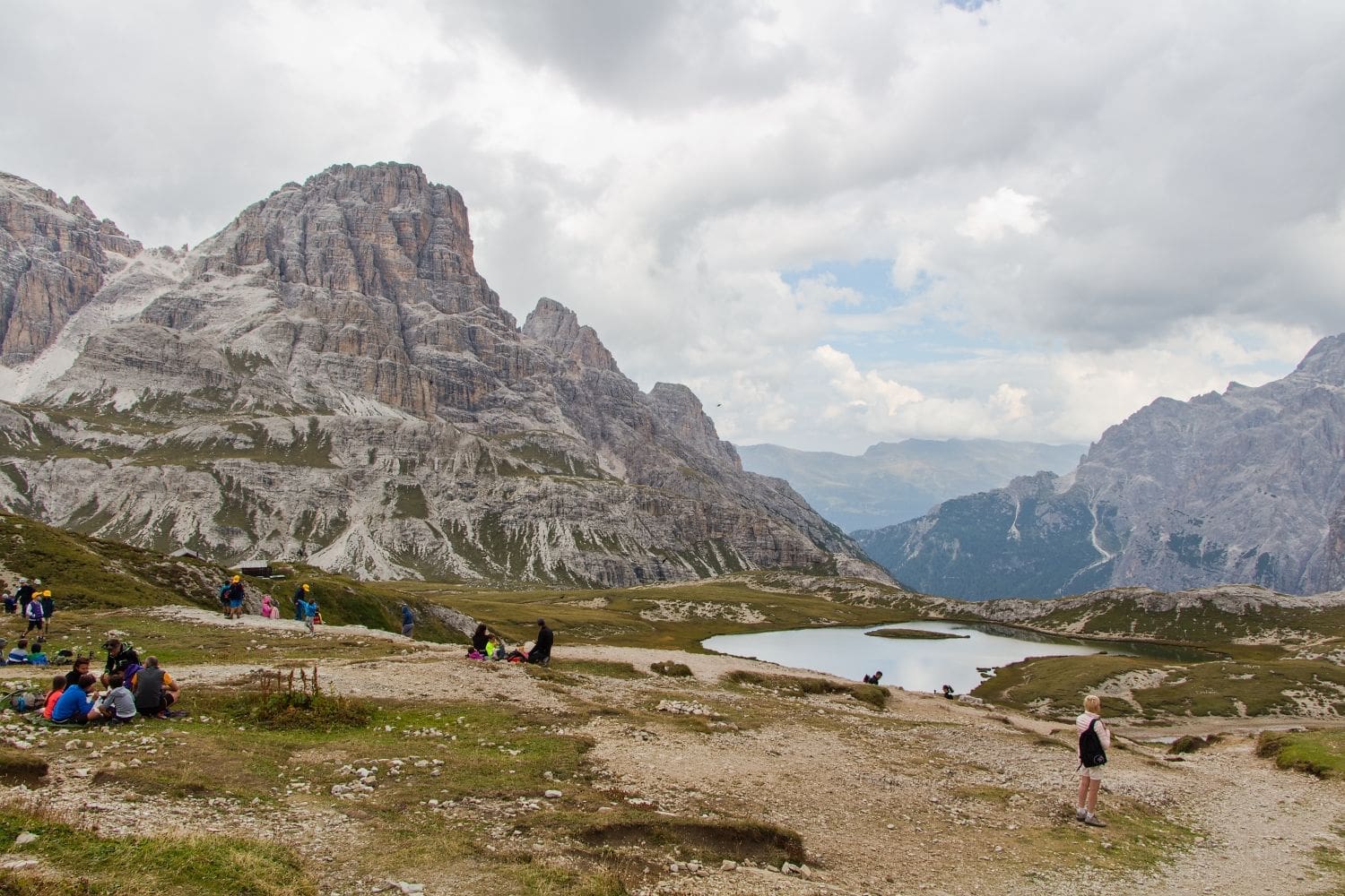 People are scattered around a scenic mountainous landscape with rugged peaks. Some are sitting, and others are walking along a dirt path. In the background, there's a small lake reflecting the cloudy sky. Patches of grass and rocky terrain dominate the foreground.