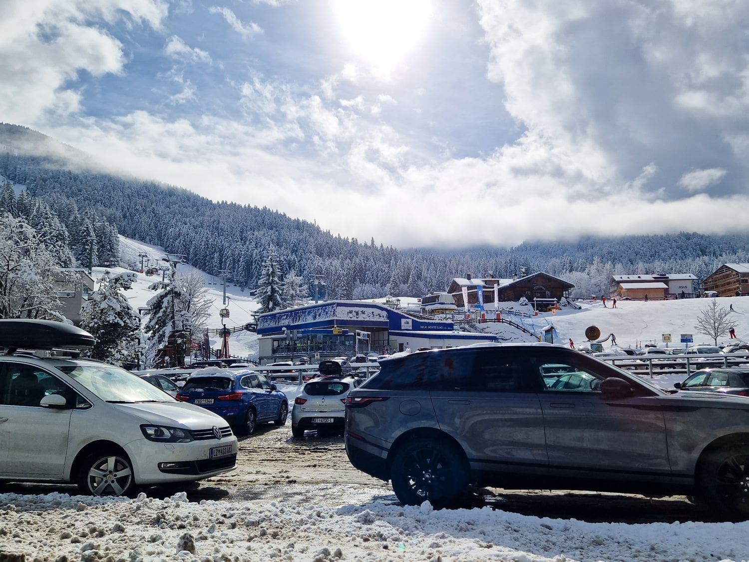 A snowy mountain landscape with a parking lot in the foreground filled with cars. The sun shines brightly through a partly cloudy sky, illuminating the ski resort buildings and snow-covered trees in the background. Ski lifts are visible ascending the mountain.