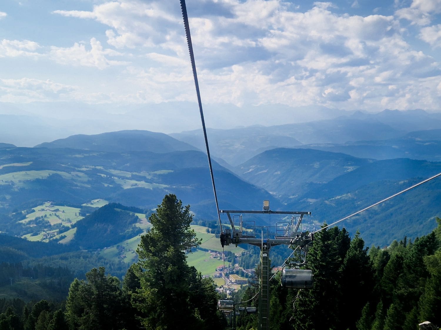 A scenic view of a mountainous landscape with valleys and hills covered in trees. In the foreground, a cable car and its supporting structure are seen ascending through the green forest. The sky is partly cloudy, and the distant mountains are slightly misty.