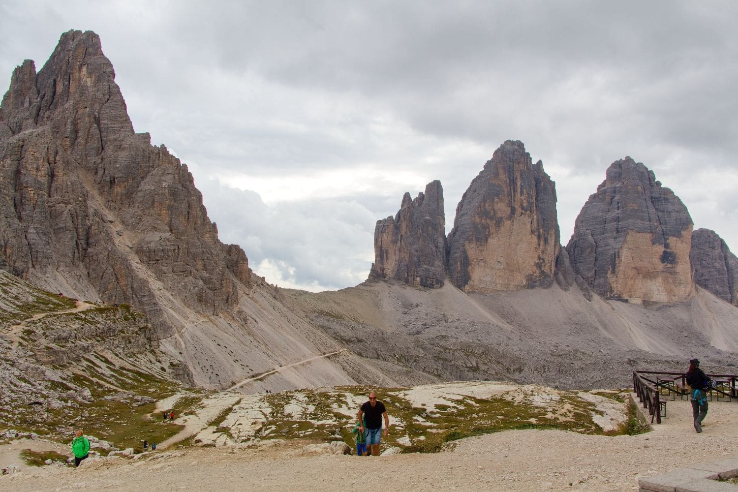 Hikers traverse a rocky, mountainous landscape under a cloudy sky, with jagged peaks in the background. The scene is dominated by gray and brown tones, with sparse vegetation. A wooden railing and viewing platform are visible on the right side.